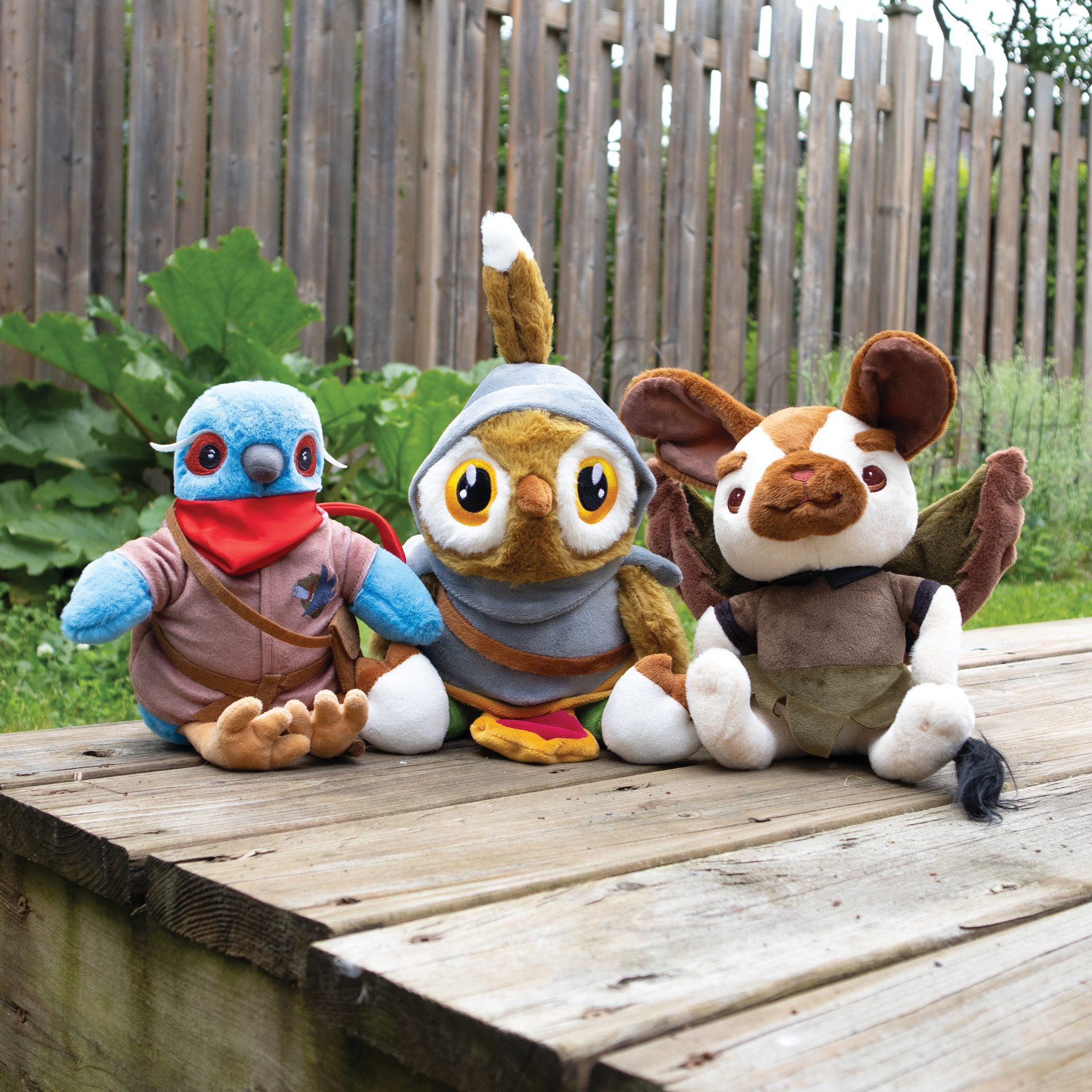 Three plush toys resembling an owl, bird, and a mouse sitting on a wooden deck with a garden background.