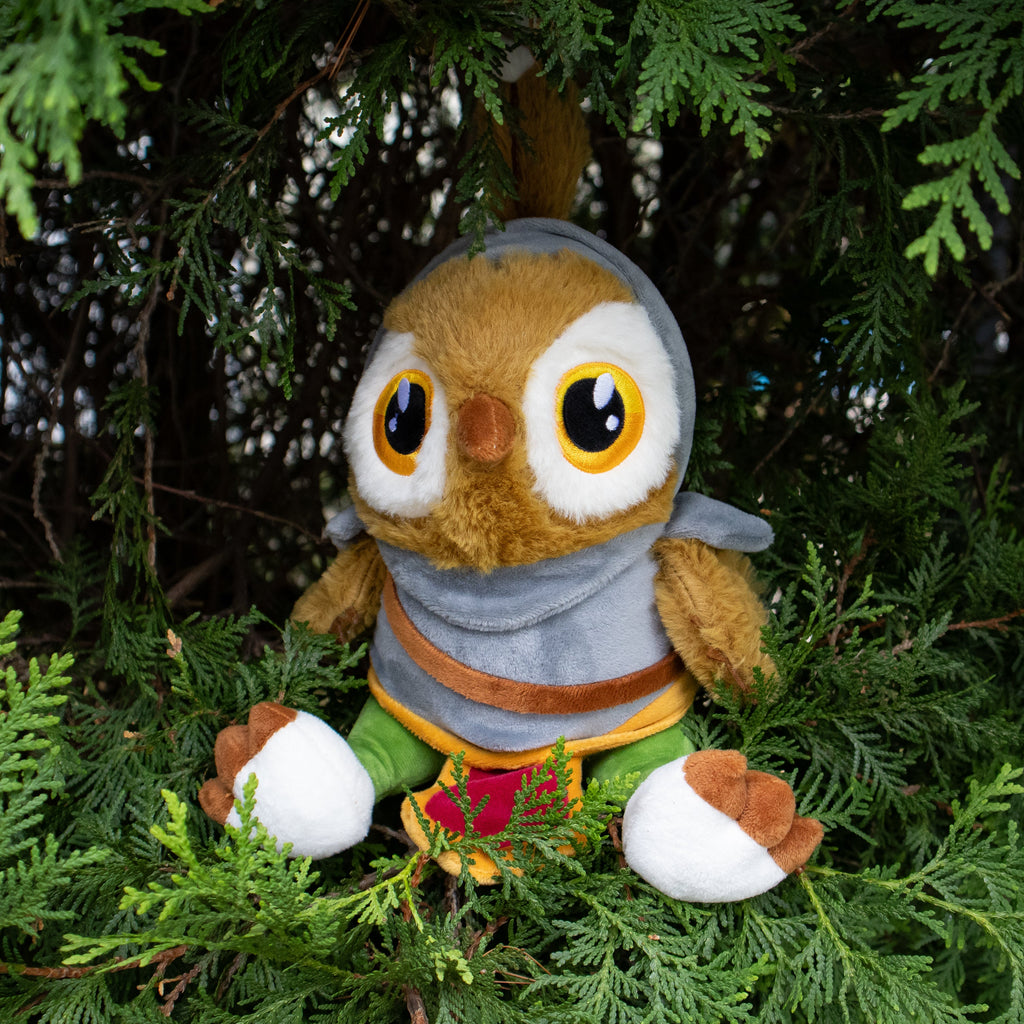 Plush owl toy with a gray hood and colorful scarf sitting among green foliage.