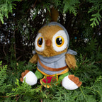 Plush owl toy with a gray hood and colorful scarf sitting among green foliage.