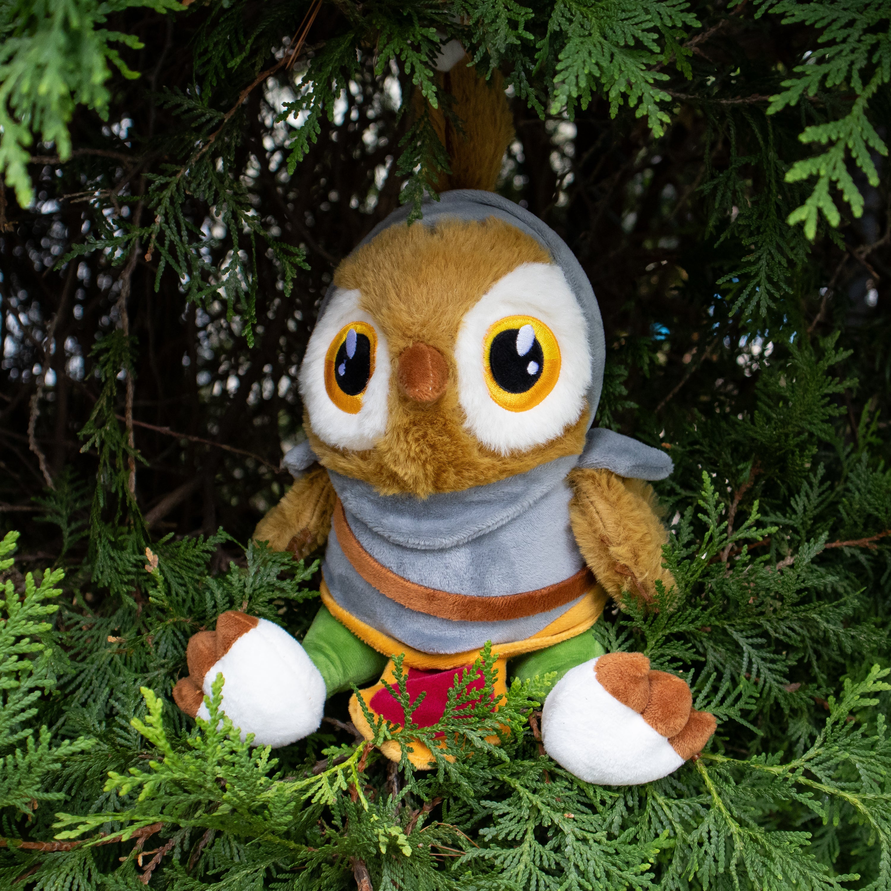 Plush owl toy with a gray hood and colorful scarf sitting among green foliage.